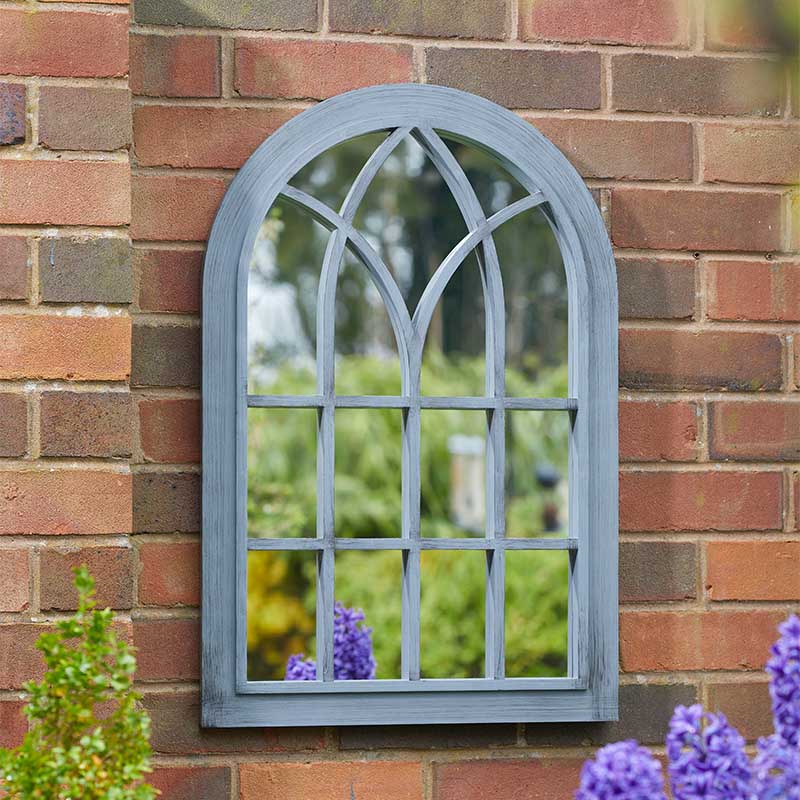 Victorian slate garden mirror with arched top and 12-pane window design mounted on red brick wall, reflecting green garden and purple flowers