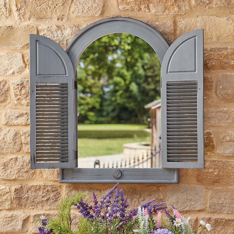 Slate grey louvre garden mirror with arched top and hinged wooden shutters mounted on stone wall, reflecting green garden and trees, with purple flowers and plants below