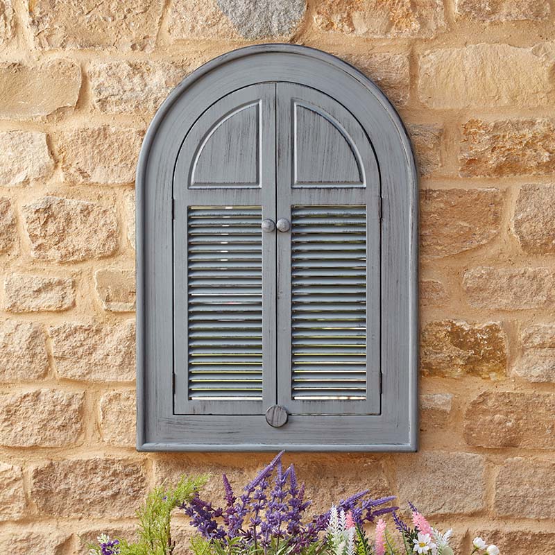 Slate grey arched louvre window mirror mounted on honey-coloured stone wall with purple and white flowering plants below