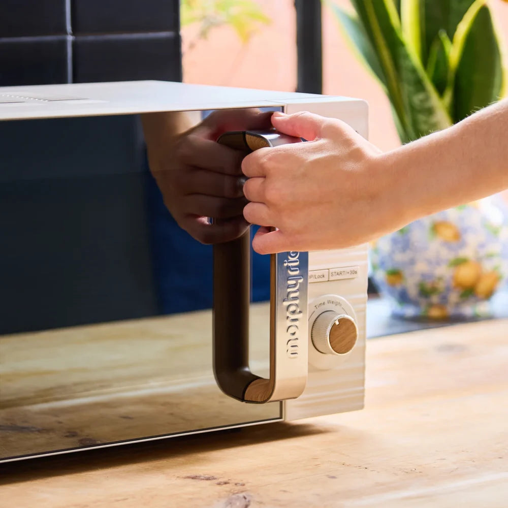 Hand opening the door of a Morphy Richards Clay white microwave with wooden accents and control dial, positioned on a wooden surface with potted plant and windows in background