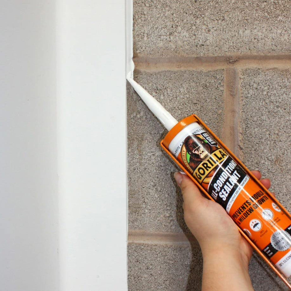 Hand holding Gorilla All Conditions Sealant 295ml white caulk tube with orange and black label against white trim and gray textured wall surface