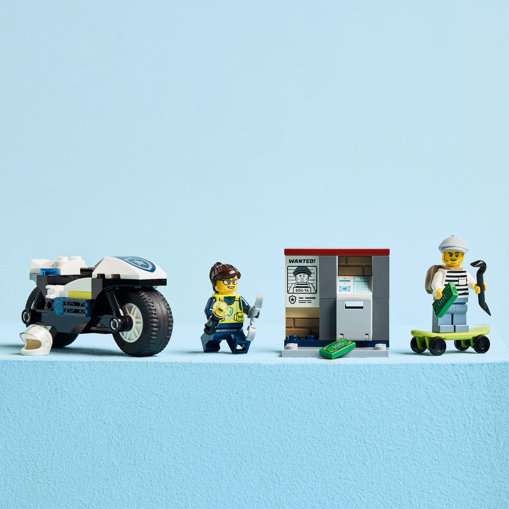 LEGO Police Motorcycle Chase set 60455 featuring a blue and white police motorcycle, two minifigures in police uniforms, a bank building with wanted poster and ATM, green currency elements, and a yellow scooter arranged on a light blue background