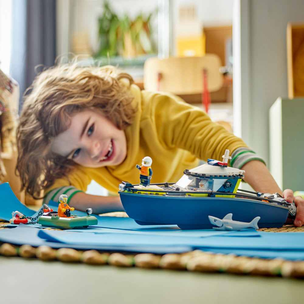 Young child in yellow shirt playing with LEGO Police Boat Chase set 60456, featuring a blue and yellow police boat with minifigures and accessories on blue play mat with rope border in bright playroom setting