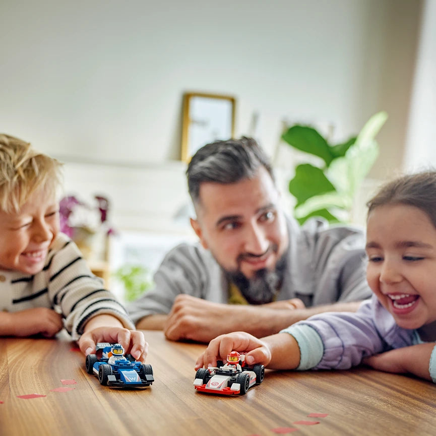 Man and two young children playing with blue and red LEGO F1 race cars on a wooden table in a bright living room with green plants and wooden framed mirror in background
