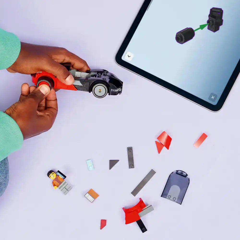 Hand holding red and black LEGO EV Supercar model with disassembled components displayed below including minifigure, red chassis pieces, gray technical parts, and blue backpack, with tablet showing digital assembly instructions in background