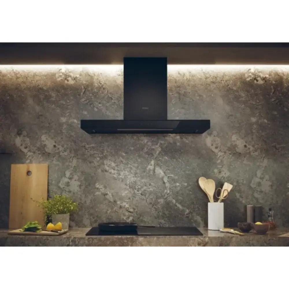 Black Haier chimney cooker hood 90cm mounted on textured grey concrete kitchen backsplash with illuminated overhead lighting, stainless steel stovetop, wooden cutting board, potted herbs and kitchen utensils visible on countertop