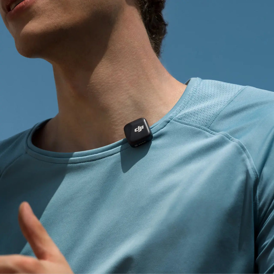 Man wearing light blue t-shirt with DJI Mic Mini wireless microphone transmitter clipped to chest against clear blue sky background