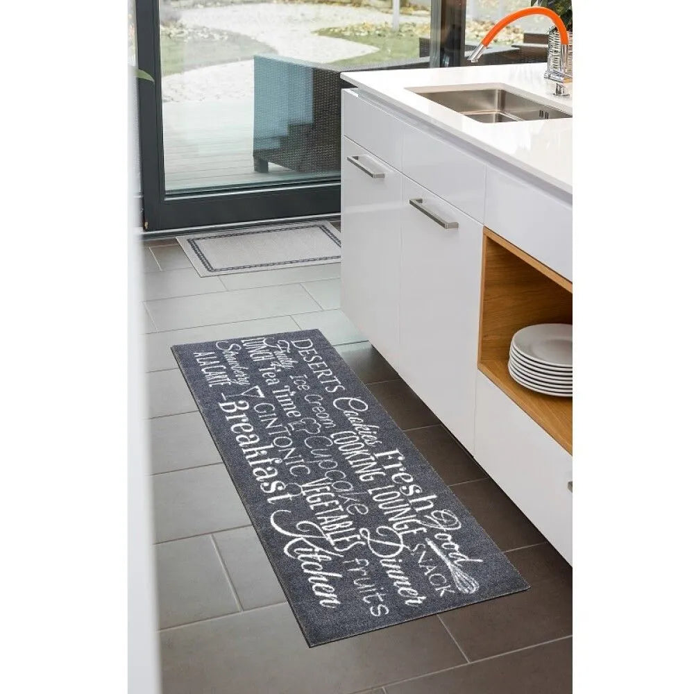 Dark grey kitchen runner mat with white decorative text and swirling script patterns, positioned in front of white cabinetry and modern kitchen island with wooden shelving, light tiled flooring visible with glass doors to outdoor patio in background