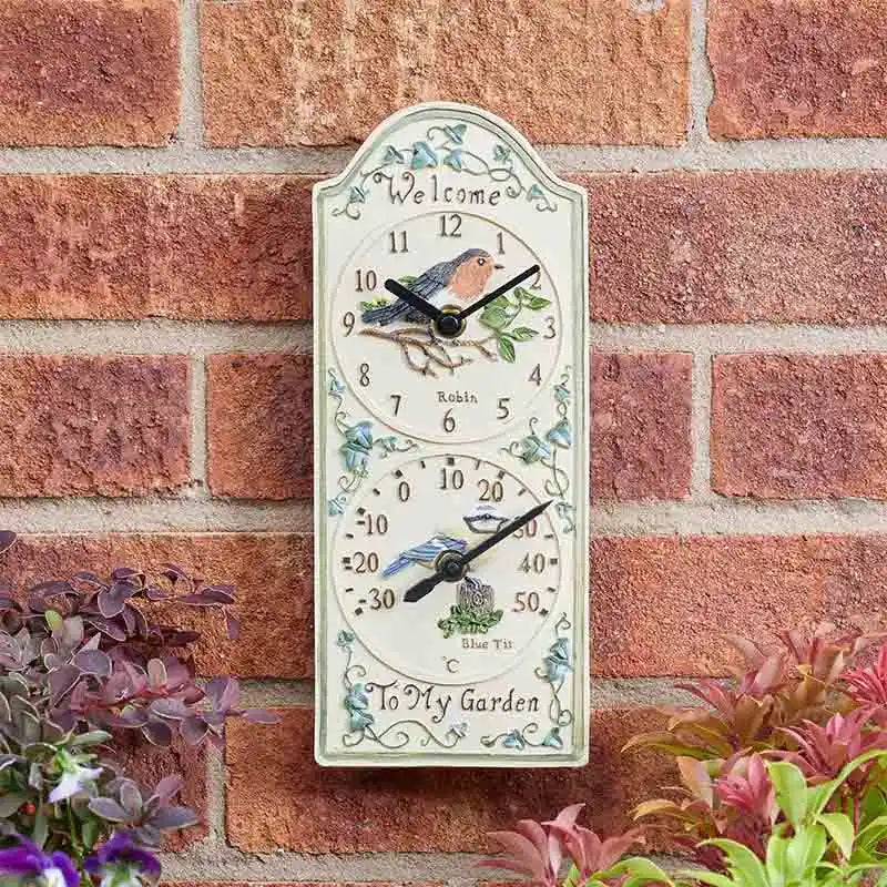Cream ceramic garden wall clock with robin bird design and integrated thermometer mounted on red brick wall with purple and pink flowers, featuring decorative leaf motifs and "Welcome to My Garden" text