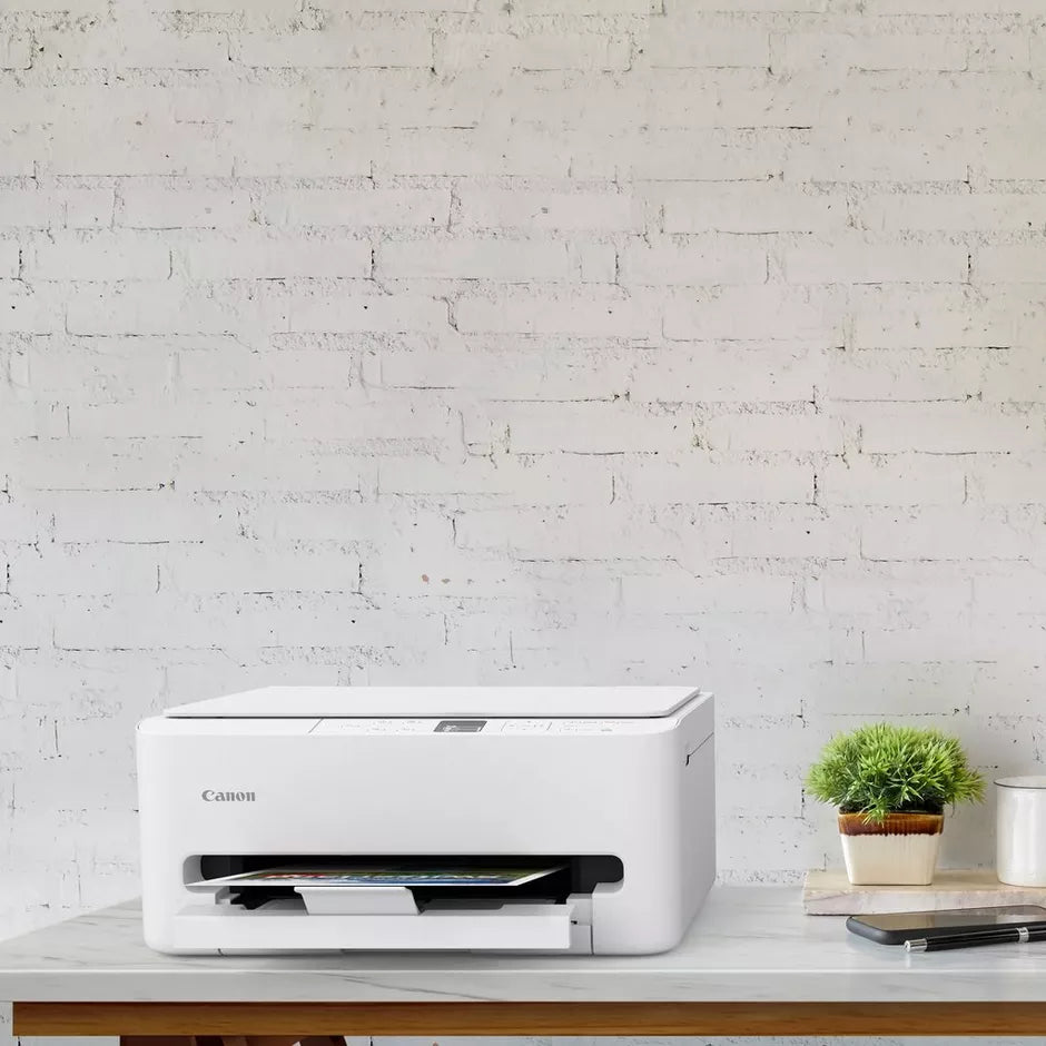 Canon TS6550i white wireless inkjet printer on wooden desk against white brick wall with small potted plant and notebook
