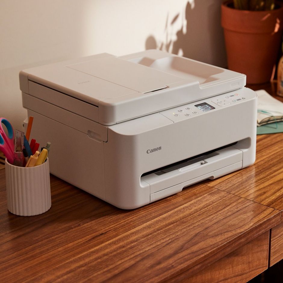 White Canon TS6550i all-in-one wireless inkjet printer on wooden desk next to pink pen holder and potted plant, with flatbed scanner lid open and control panel visible