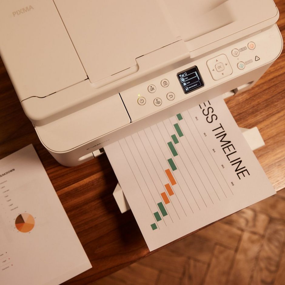 Canon PIXMA TS6550i white wireless inkjet printer with control panel displaying settings, printing a timeline chart with green and orange bars on wooden surface surrounded by printed documents with pie charts