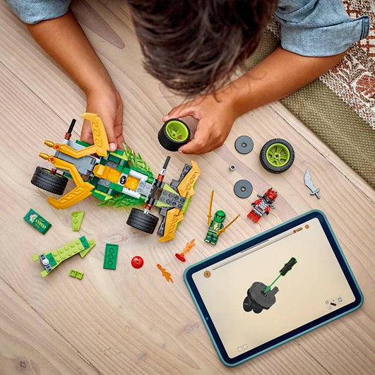 Child assembling green and yellow Lego pull-back race car with wheels and minifigure on wooden floor with instruction tablet and scattered Lego pieces