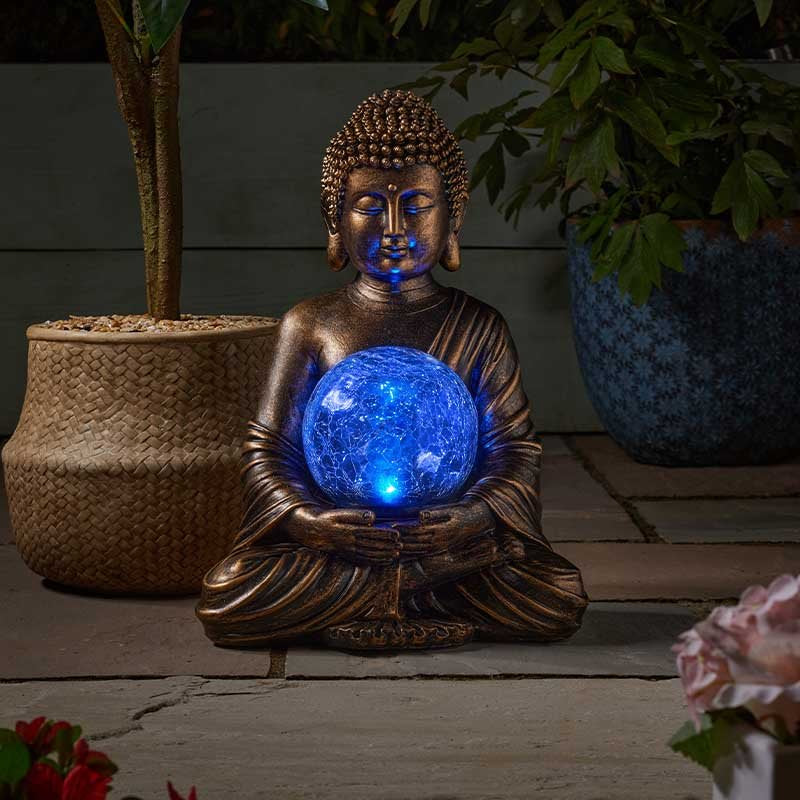 Bronze Buddha statue holding illuminated blue glowing orb sphere, seated in meditation pose surrounded by potted plants and flowers on wooden deck garden setting