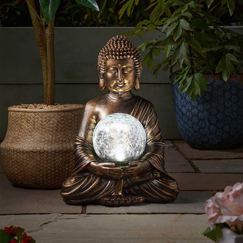 Bronze Buddha statue holding illuminated crackle glass solar light sphere, seated in meditation pose surrounded by potted plants and flowers in a garden setting