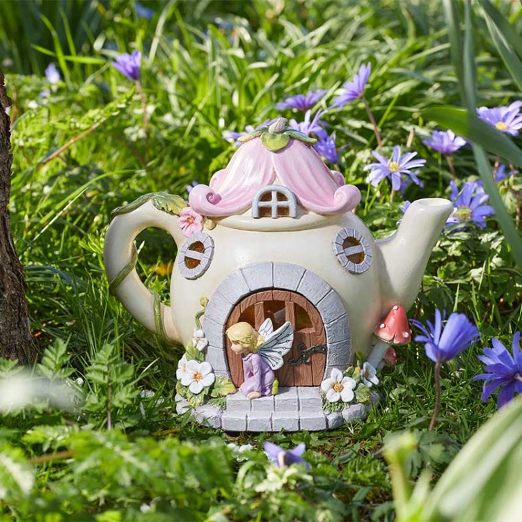 Whimsical fairy teapot garden ornament with pink roof and purple door nestled among purple flowers and green grass, featuring solar light functionality