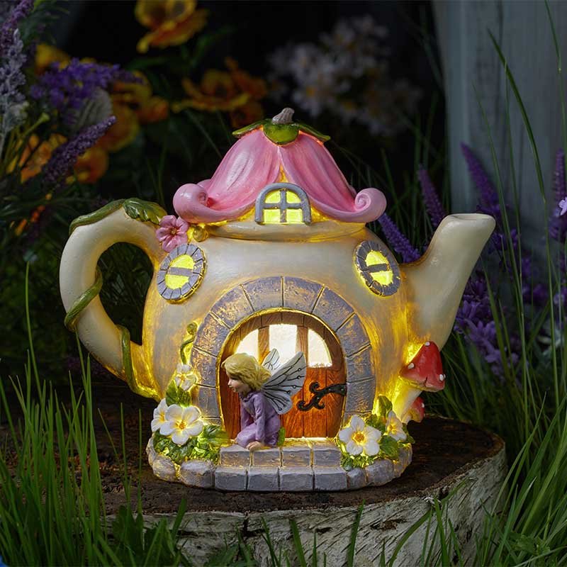 Whimsical fairy teapot solar light with pink curved roof, cream ceramic body, illuminated yellow windows and doors, decorated with colorful flowers and a tiny fairy figurine, surrounded by purple flowers and green plants in a garden setting