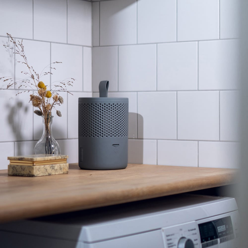 Grey cylindrical moisture absorber with perforated design and fabric handle displayed on wooden countertop next to glass vase with dried flowers, white subway tile backsplash background