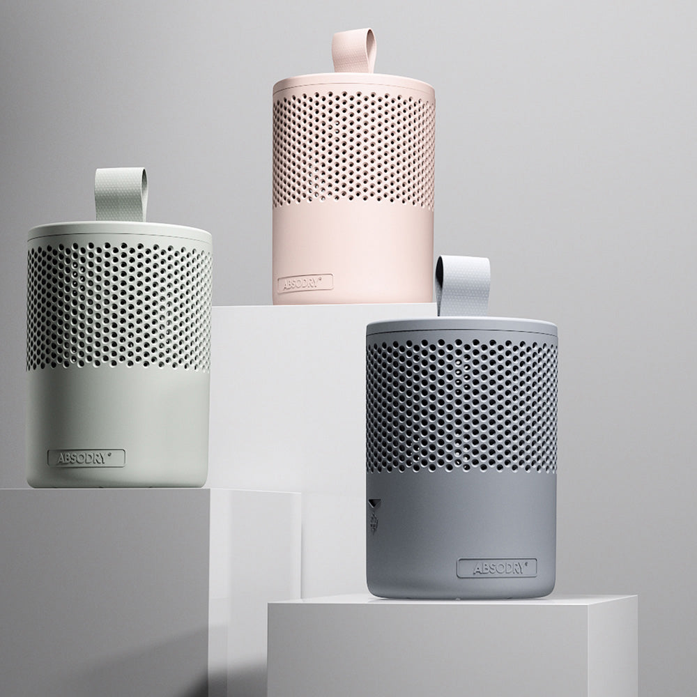Three cylindrical AbsoDry moisture absorbers in varying sizes displayed on white platforms, featuring perforated metal tops in grey, white and pink finishes with fabric carry handles