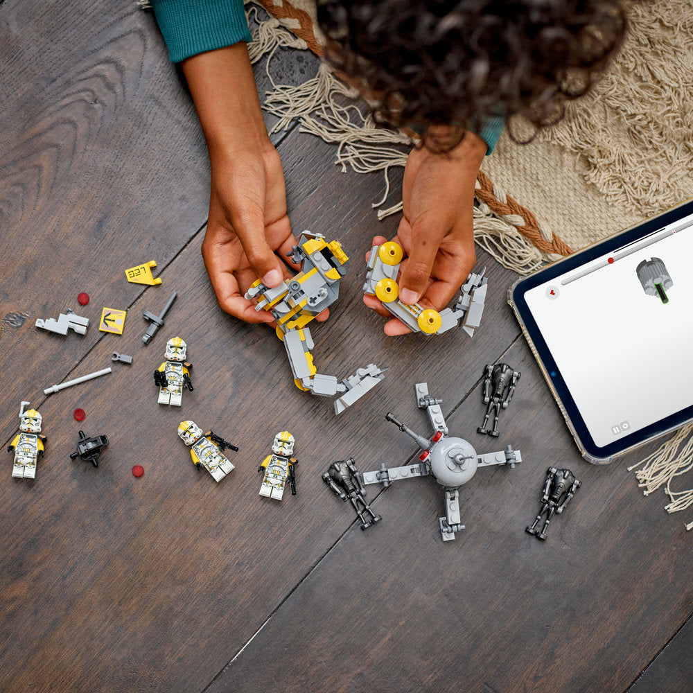 Child's hands holding yellow and white LEGO Star Wars clone trooper minifigures and buildable droids on wooden floor with instruction manual and additional black LEGO figures scattered around
