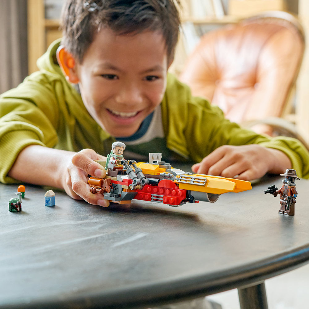 A young child in a lime green jacket smiles while playing with a LEGO Star Wars Cobb Vanth's Speeder set on a grey wooden table, featuring red and yellow building blocks, minifigures, and small accessories