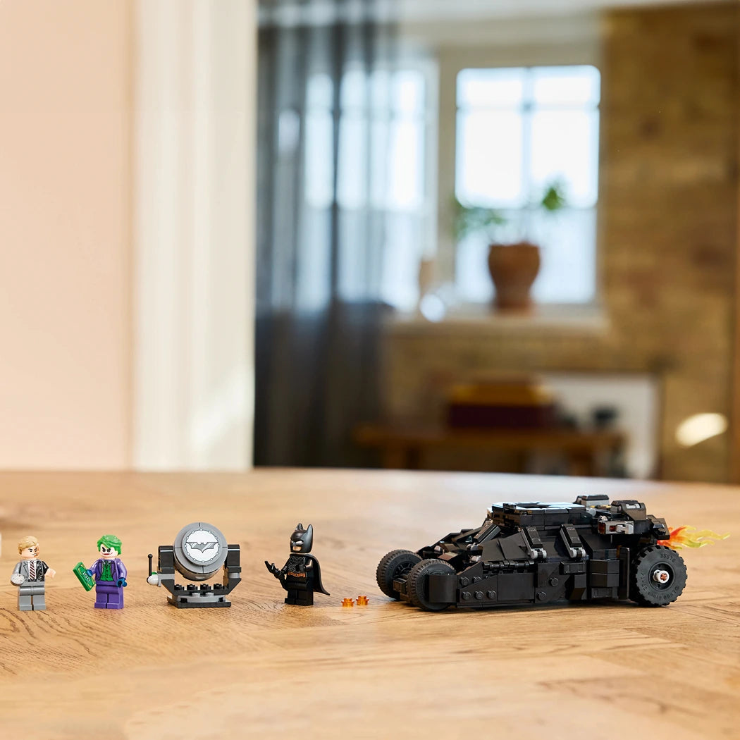 LEGO Batman set 76303 displayed on wooden table featuring black Batmobile Tumbler vehicle with flame details, Batman minifigure, Joker in purple suit, Two-Face character, and accessories including Gotham City police searchlight, arranged against blurred home interior background with window and curtains