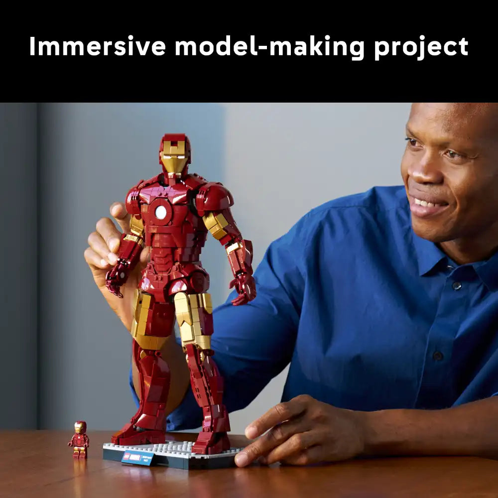 Man in blue shirt holding large red and gold LEGO Iron Man Mark 3 buildable figure with detailed armor plating, standing next to smaller minifigure on table during immersive model-making project
