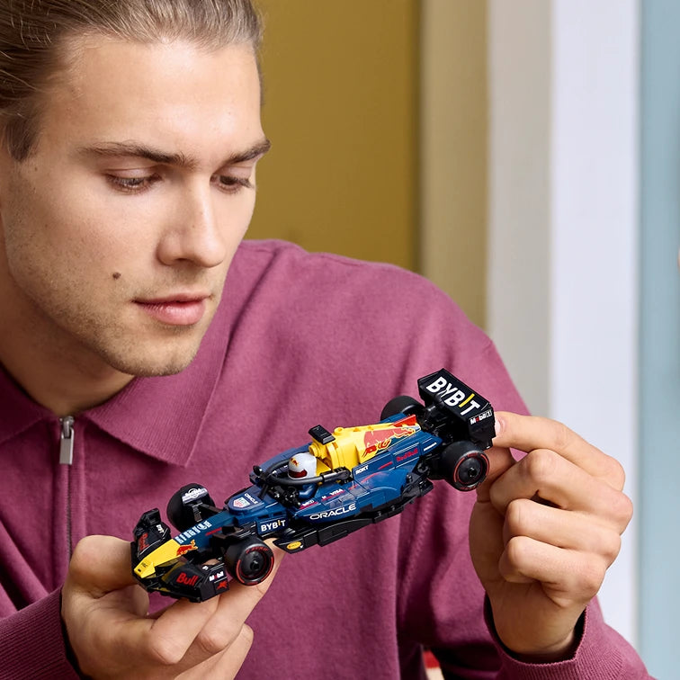 Man in burgundy polo shirt holding LEGO Oracle Red Bull Racing RB20 F1 race car model featuring blue and yellow livery with detailed minifigure driver against neutral interior background