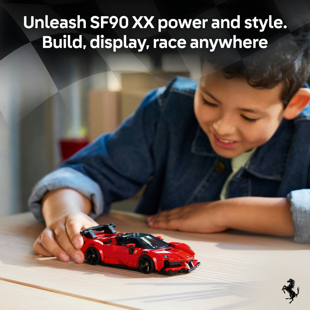 Young boy in blue jacket playing with red LEGO Ferrari SF90 XX sports car model on wooden table, showcasing buildable toy car with black accents and wheels