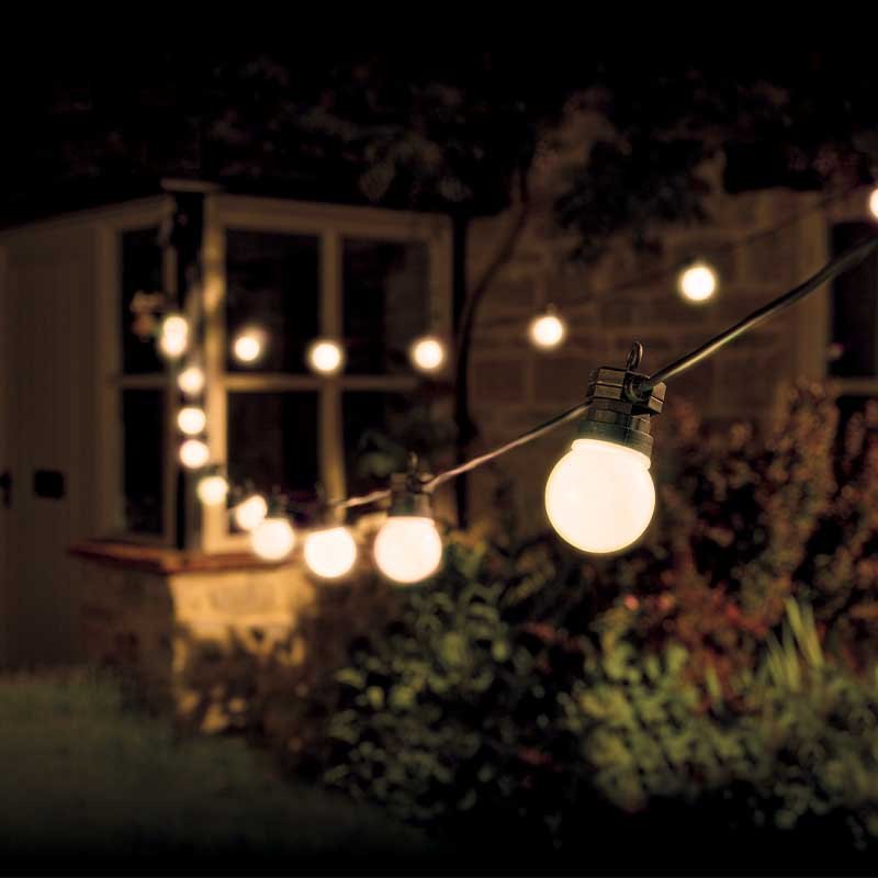String of 20 warm white LED festoon lights strung across an outdoor garden at night, illuminating plants and a modern house with lit windows in the background