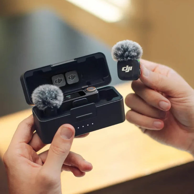 Hands holding DJI Mic 3 wireless microphone system showing two black transmitter units with gray windscreen covers and one receiver unit inside black charging case with LED battery indicator lights