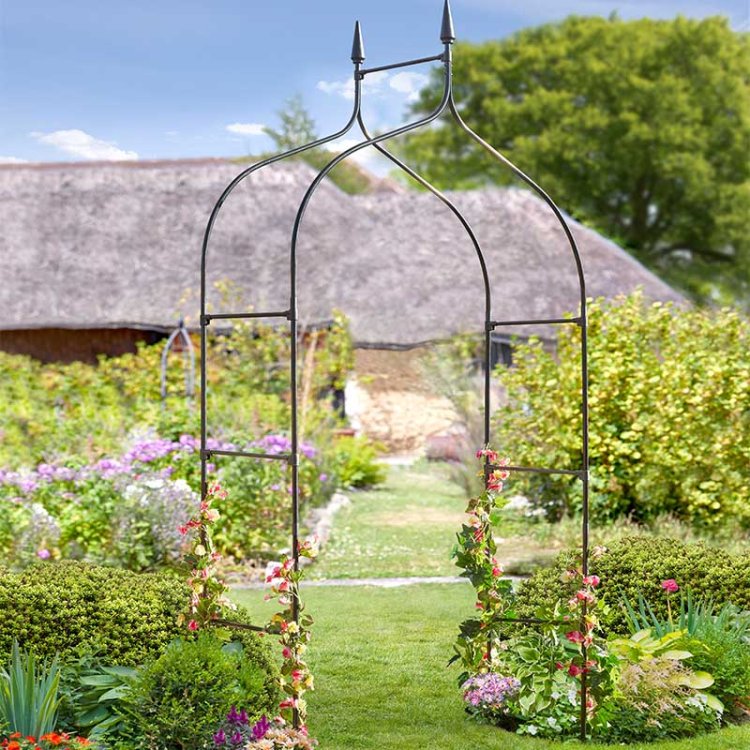 Black metal garden arch with decorative finials in a lush cottage garden setting, surrounded by purple flowers, green hedging and climbing plants, with a thatched roof cottage visible in the background
