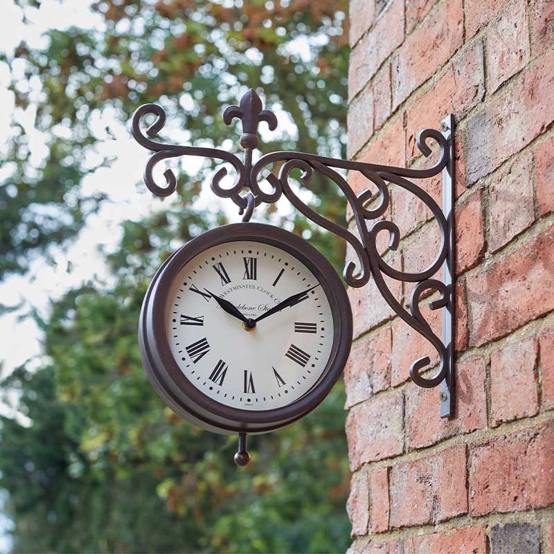 Ornate dark brown wrought iron garden wall clock with Roman numerals mounted on a red brick wall, featuring decorative scrollwork bracket and an 8-inch cream dial face with visible time display, with blurred green foliage in the background