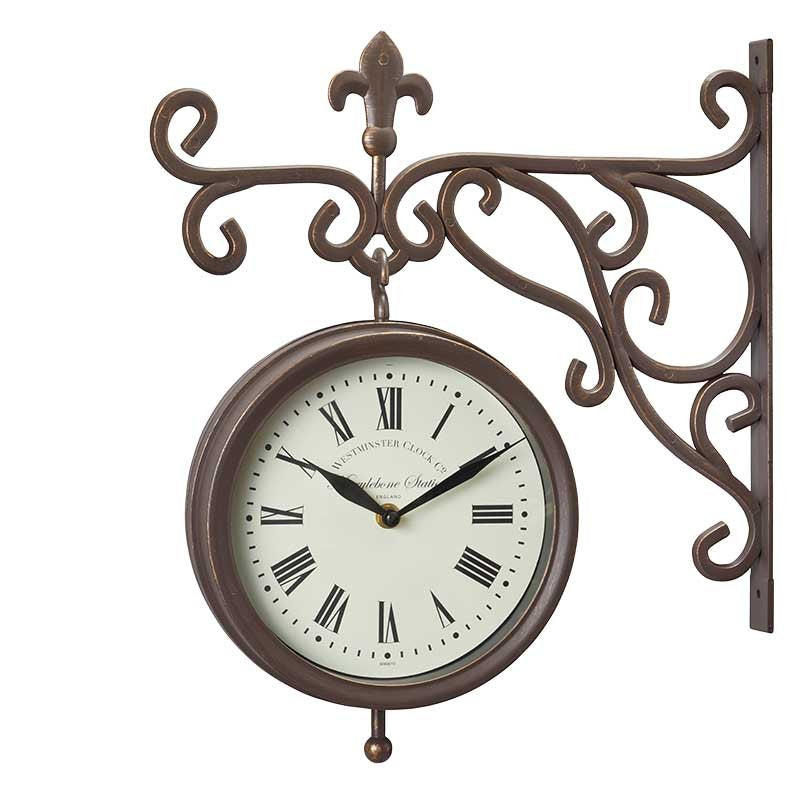 Vintage-style bronze wall-mounted clock with ornate scrollwork bracket, cream face with Roman numerals, and decorative fleur-de-lis finial, Marylebone Station design
