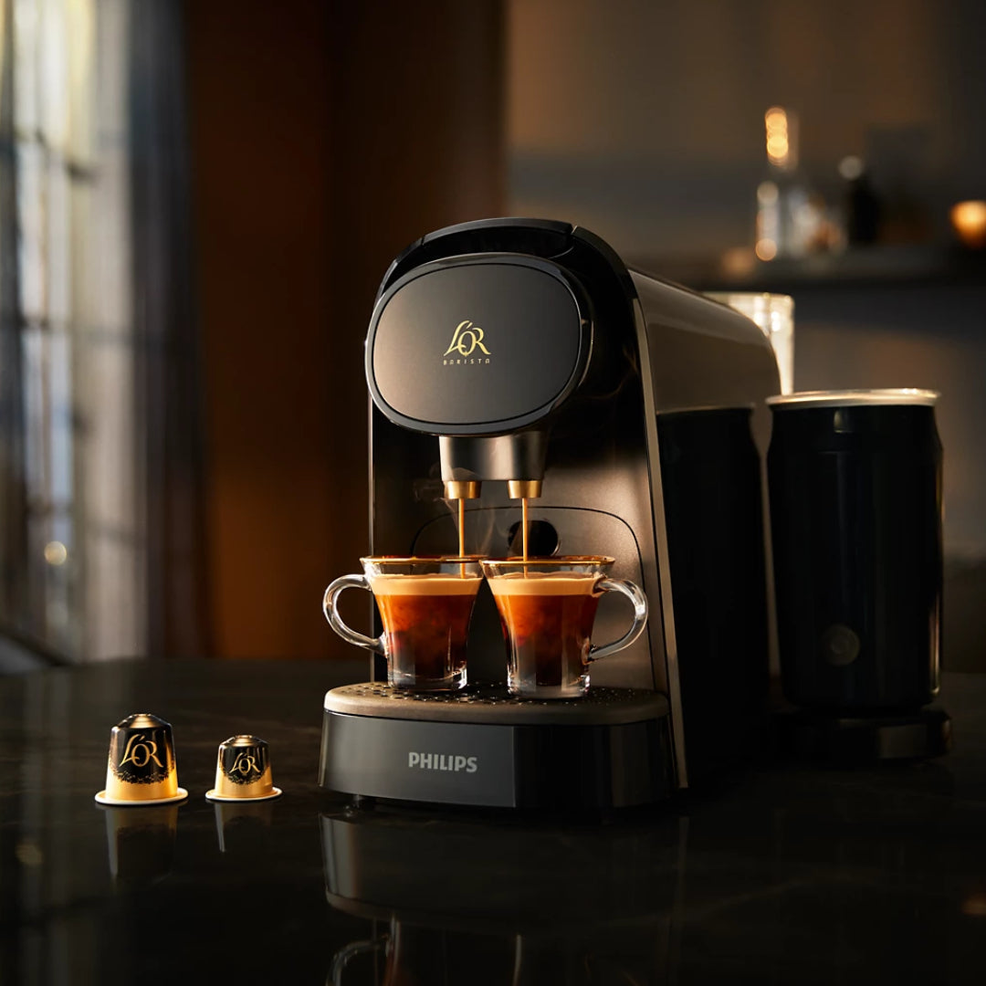 Philips L'OR Barista deep black capsule coffee machine dispensing espresso into two glass cups with gold-rimmed L'OR capsules displayed in front, against a blurred warm-lit background