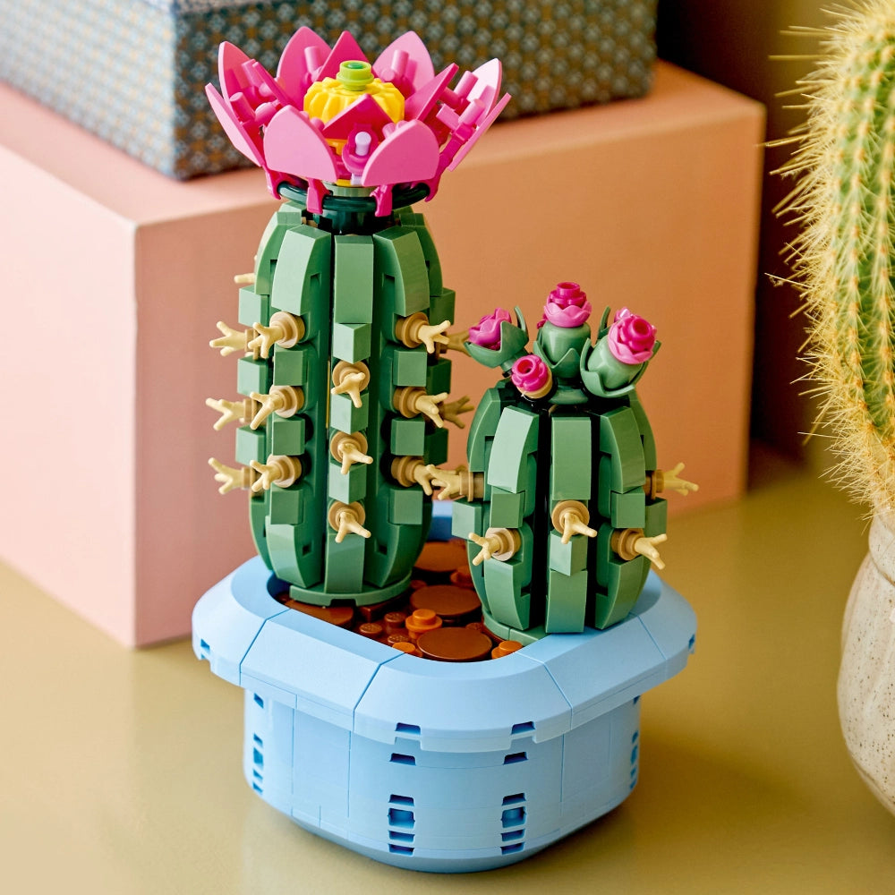 LEGO Flowering Cactus set 10309 featuring two buildable cacti with pink blooms and tan spines in a light blue pot with brown soil, displayed on a shelf with colorful home decor in the background