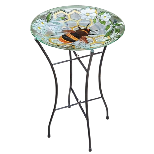 Glass bird bath with painted bee design on round top, supported by black metal stand with curved legs, featuring white flowers and green foliage, ideal for garden decoration