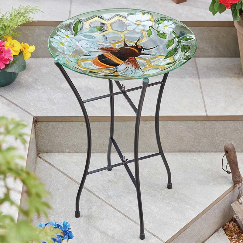 Glass bird bath with hand-painted bee and white flowers design on black metal stand in outdoor garden setting surrounded by potted flowers