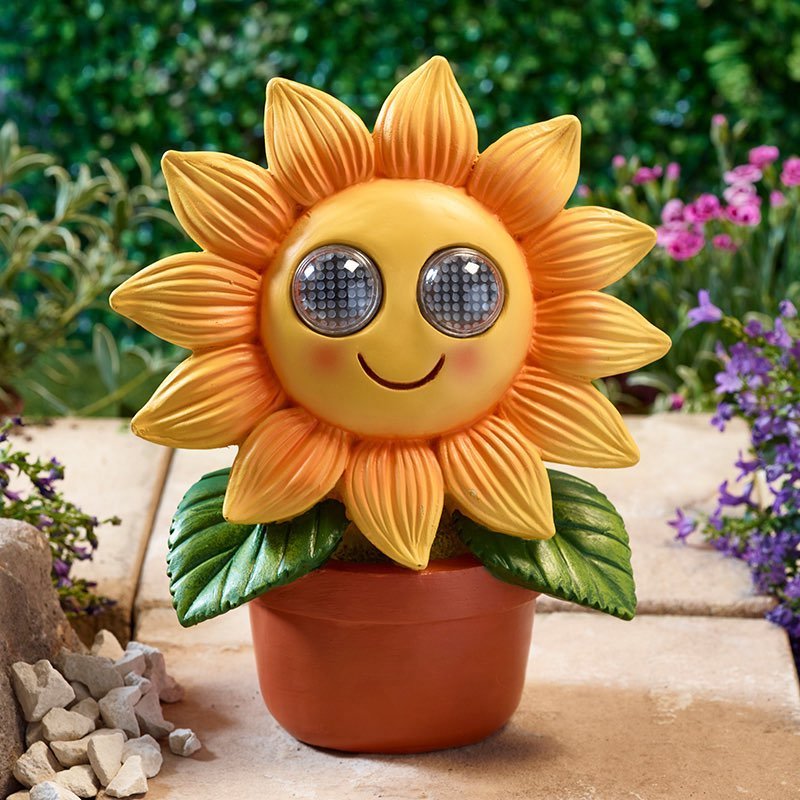 Cheerful yellow and orange solar-powered sunflower ornament with large round eyes in terracotta pot, displayed in garden setting with purple flowers and green foliage
