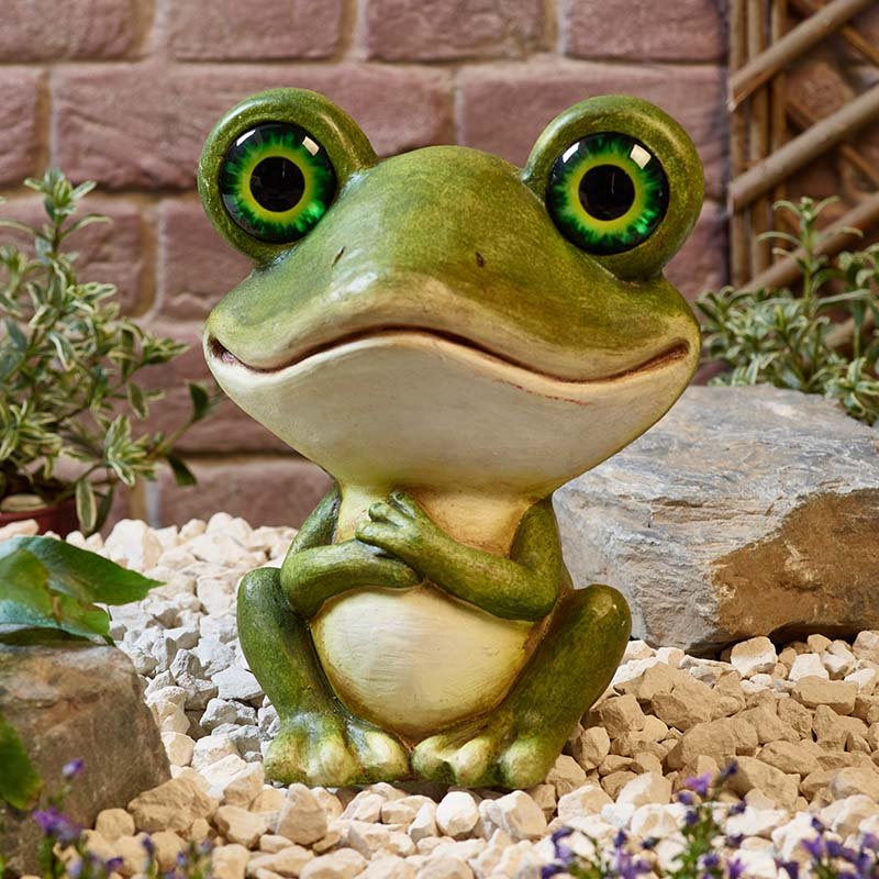 Green and cream resin garden frog ornament with large bright yellow-green eyes and cheerful expression, positioned on white gravel surrounded by small purple flowers and green foliage against a brick wall background