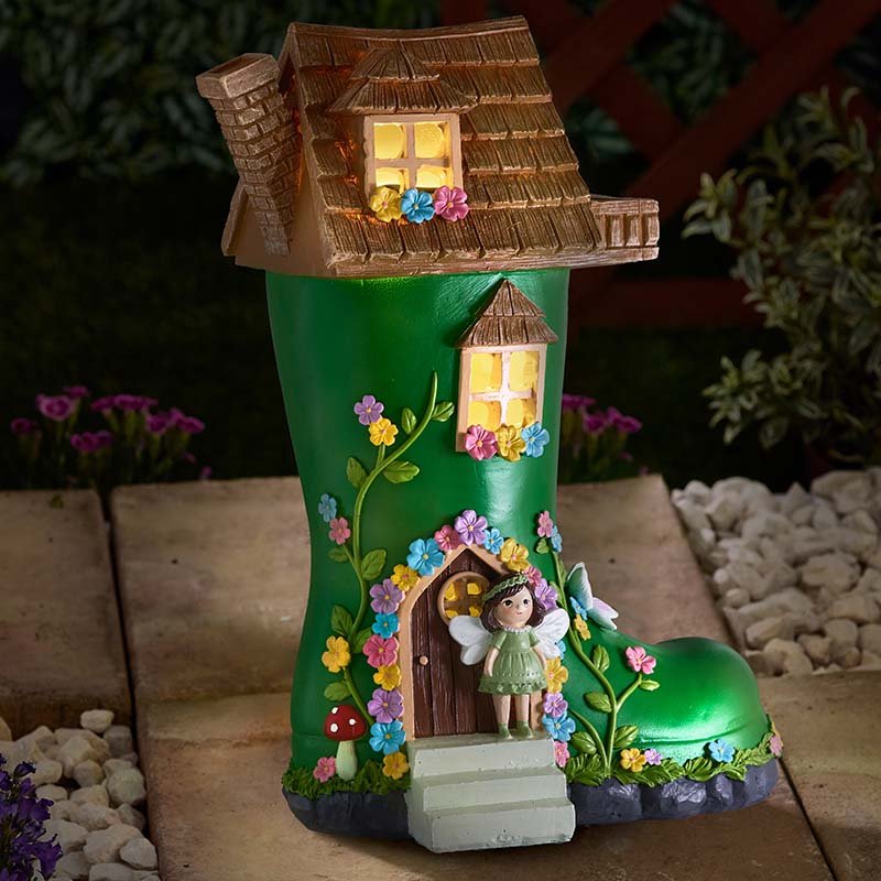 Green wellington boot shaped garden ornament with brown thatched roof, illuminated windows, colorful painted flowers, fairy figurine at entrance, and solar-powered lighting feature displayed on outdoor patio with plants in background