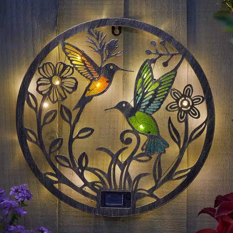 Solar-powered garden ornament featuring two illuminated hummingbirds with colored glass wings in amber and green, surrounded by decorative black metal flowers and foliage on a circular brass-toned metal disc, mounted on wooden fence with purple and red flowers visible