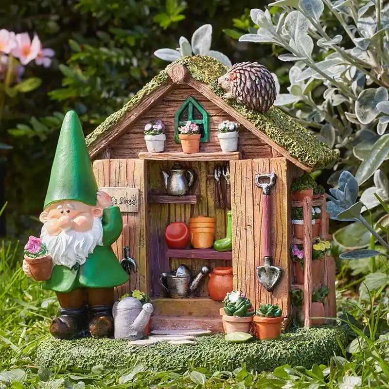 Garden gnome figurine with green pointed hat standing beside a wooden shed miniature with moss-covered roof, featuring tiny potted flowers, gardening tools, watering can and decorative plants in a natural garden setting