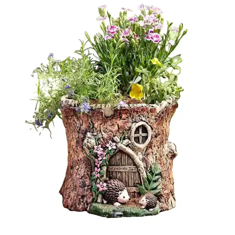 A whimsical fairy garden planter shaped like a tree trunk with a small door, window, hedgehog figures, and green plants with pink and purple flowers, shown on a plain white background.
