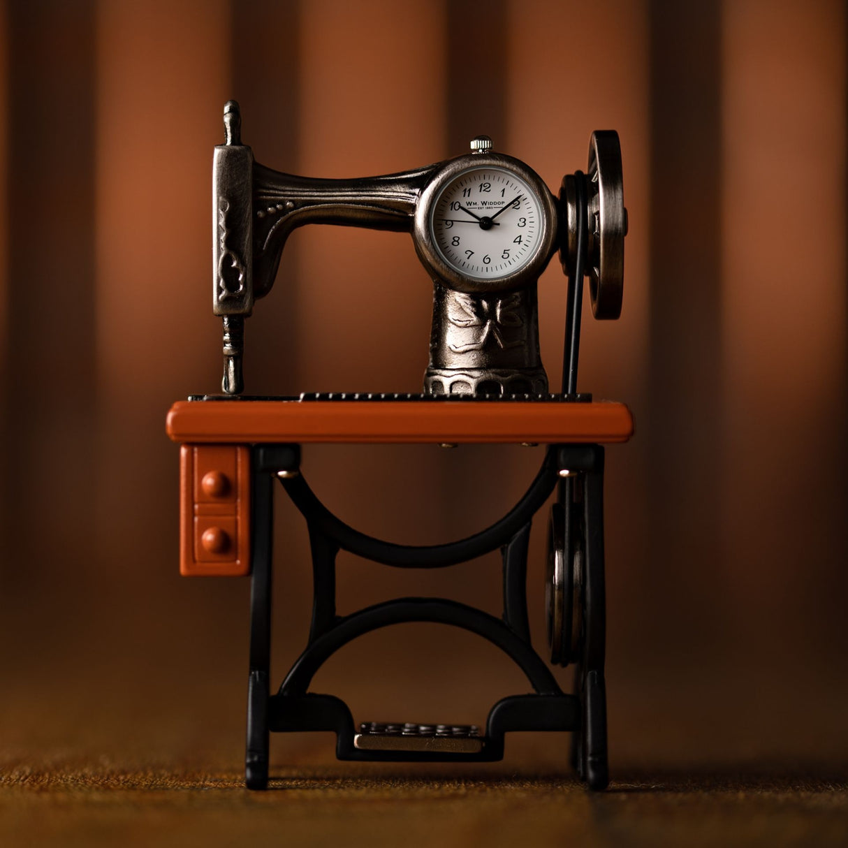 Widdop miniature sewing machine clock with cast metal body, orange wooden base and platform, white clock face showing approximately 10:10, featuring decorative details and vintage styling against a warm brown background
