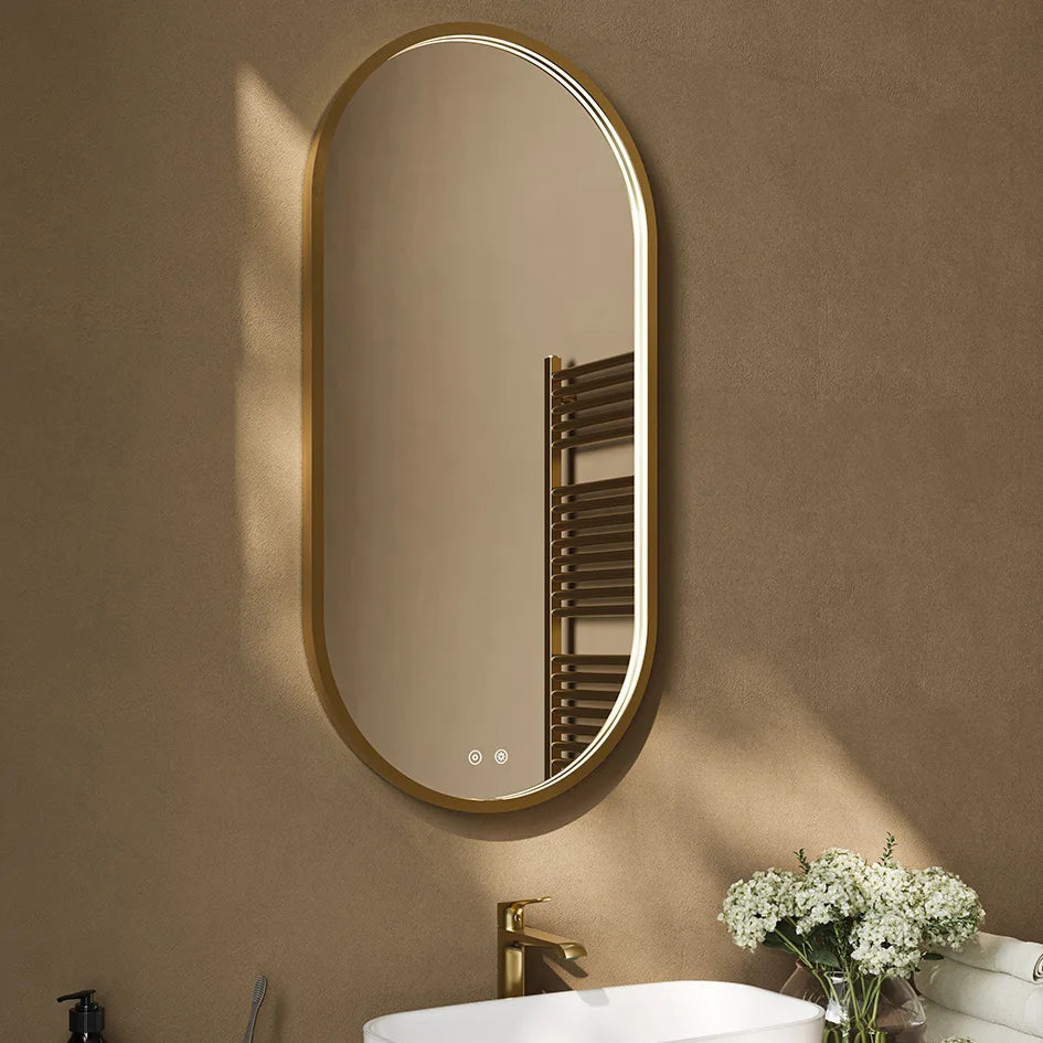 Aqualla Solaris Pill LED bathroom mirror with brushed brass frame mounted above white sink in modern bathroom setting with white flowers and brass tap visible
