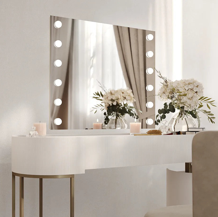 Modern bathroom vanity setup featuring a white ribbed curved desk with brass legs, illuminated LED mirror with 20 warm white bulbs around the frame, white flowers in glass vases, candles, and brushes on the countertop, with beige curtains visible in the mirror reflection, contemporary minimalist aesthetic