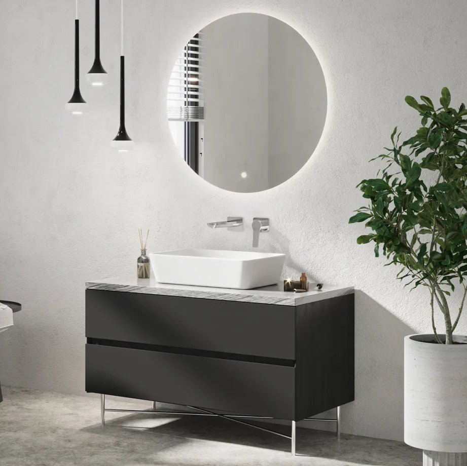 Modern bathroom vanity setup featuring a round LED-lit mirror with white illuminated frame mounted above a black floating vanity cabinet with marble countertop and white vessel sink, accompanied by chrome wall-mounted faucet, black pendant lights, and potted green plant on white concrete floor