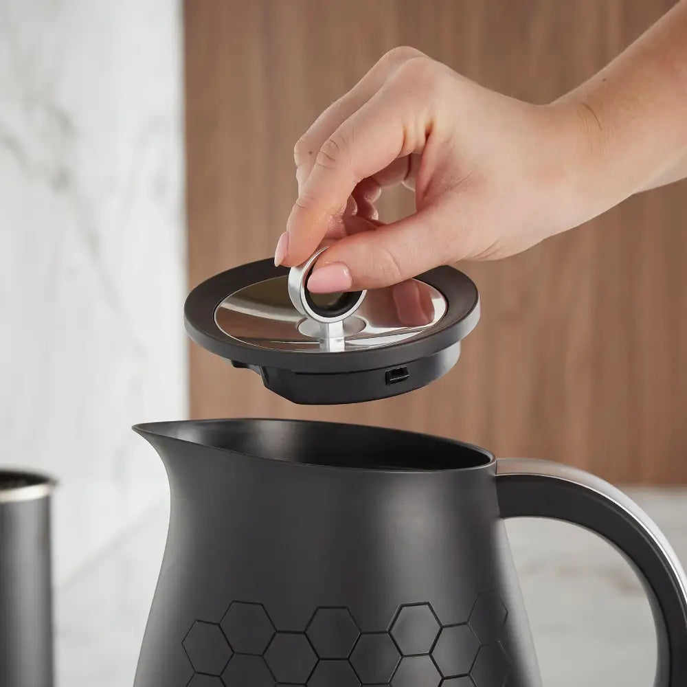Hand removing the lid from a Morphy Richards Cassini black jug kettle with hexagonal pattern design and stainless steel accents, shown against a neutral beige and white background