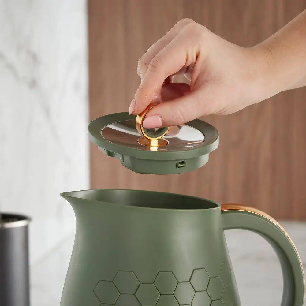Hand lifting the lid off a sage green Morphy Richards Cassini jug kettle with gold-trimmed handles and honeycomb pattern detailing, displaying the interior stainless steel lid mechanism